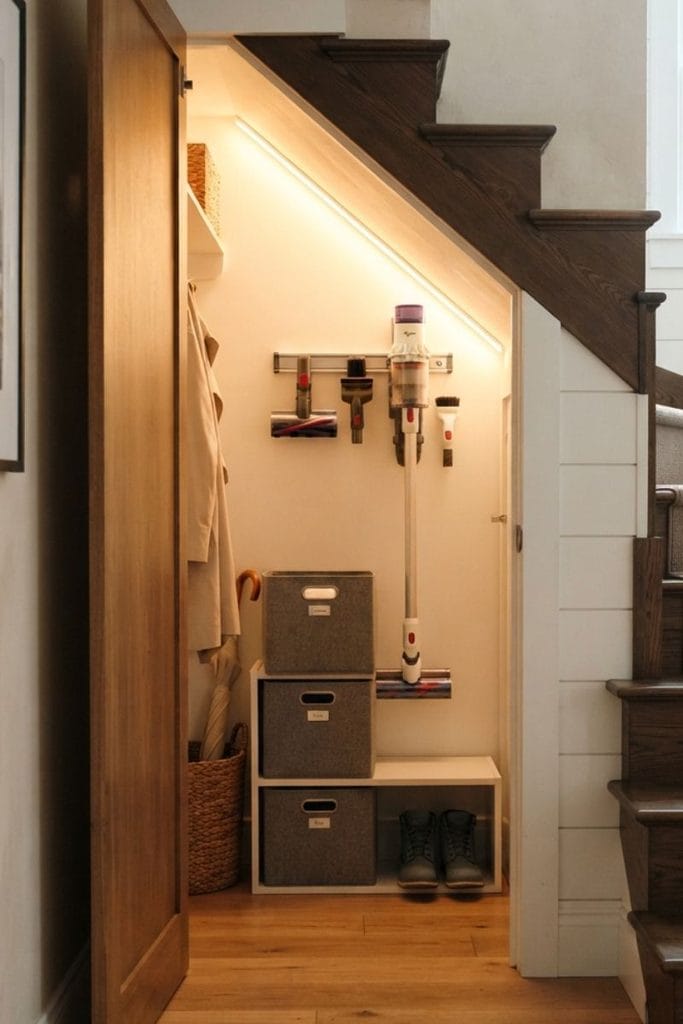 Cordless vacuum hanging inside a tidy under stairs closet with bins and a soft LED strip light
